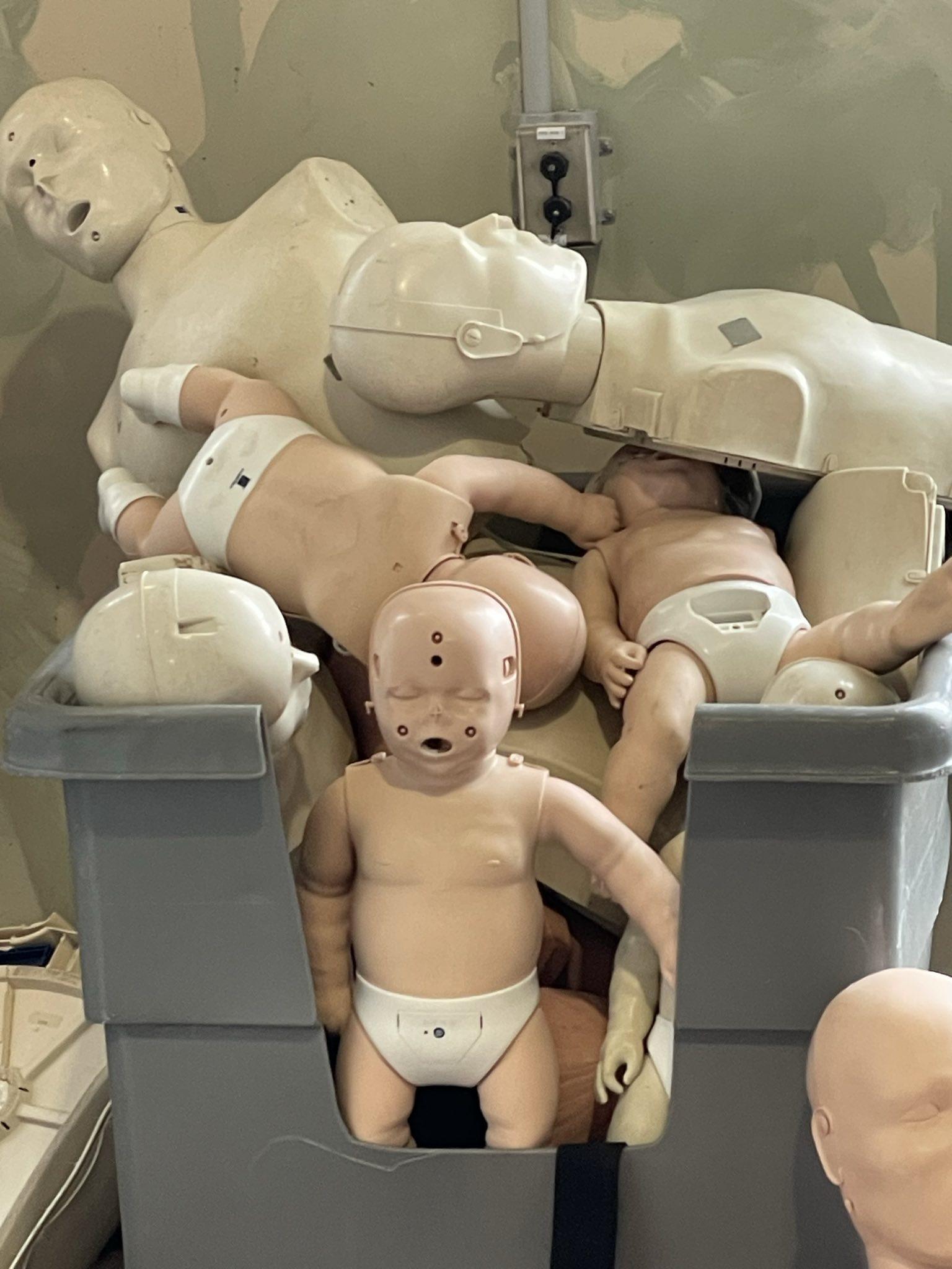 Large plastic bin full of corpselike CPR dummies, bith baby and adult. The baby is covered in holes on his face